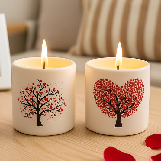 Two lit candles in cream ceramic holders decorated with red heart-shaped and tree designs, placed on a wooden table with rose petals in a cozy room setting.